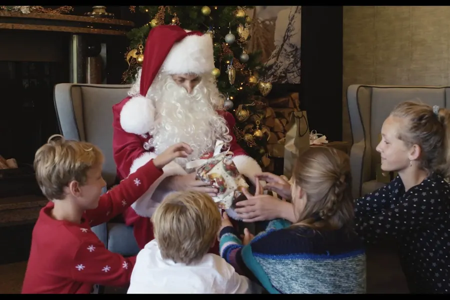 De Kerstman deelt cadeautjes uit aan kinderen bij een sfeervol versierde kerstboom.
