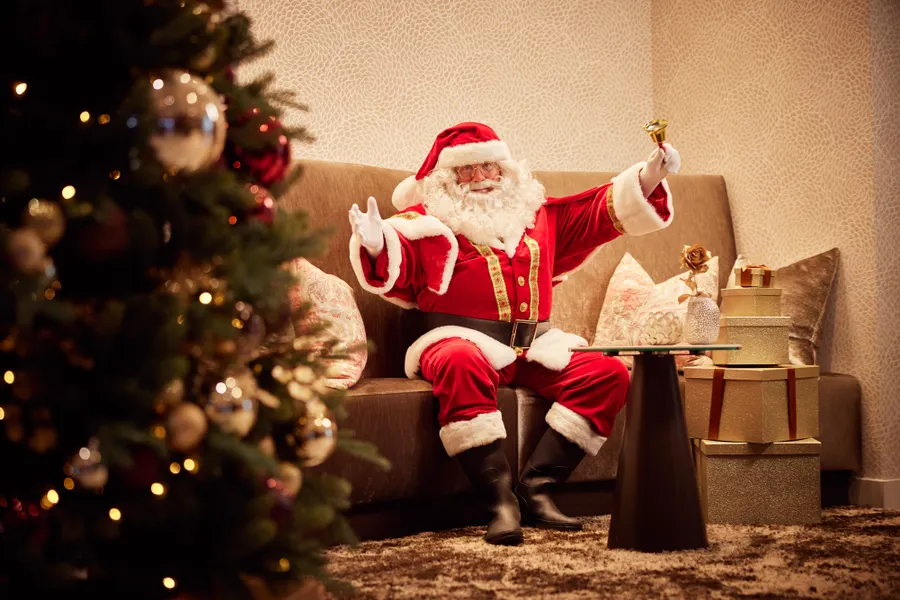 De Kerstman zit ontspannen met een cadeau naast een verlichte kerstboom in het hotel.