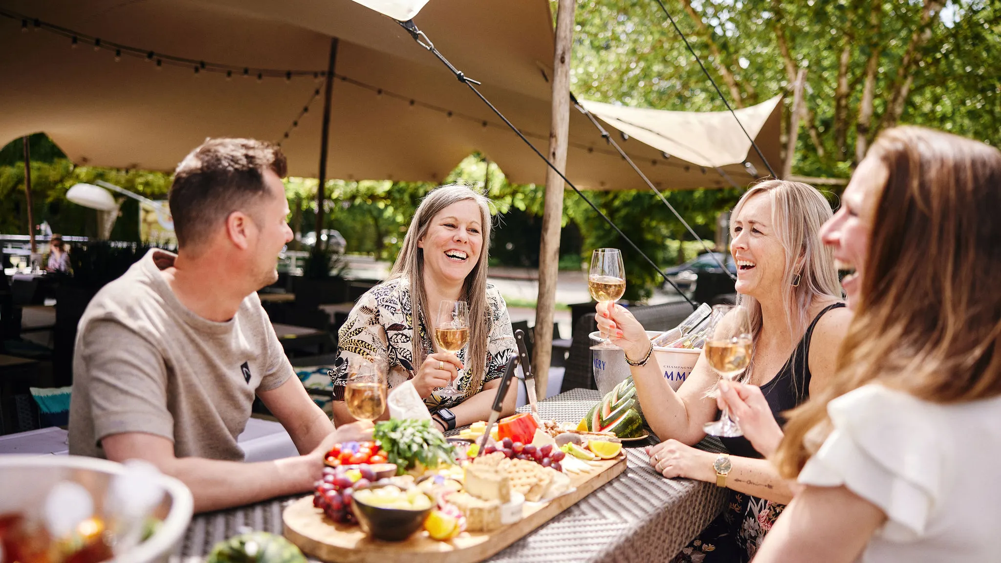 Eine Gruppe genießt gemeinsam Getränke und eine reichhaltige Wurst- und Käseplatte auf der sonnigen Außenterrasse des Hotels.