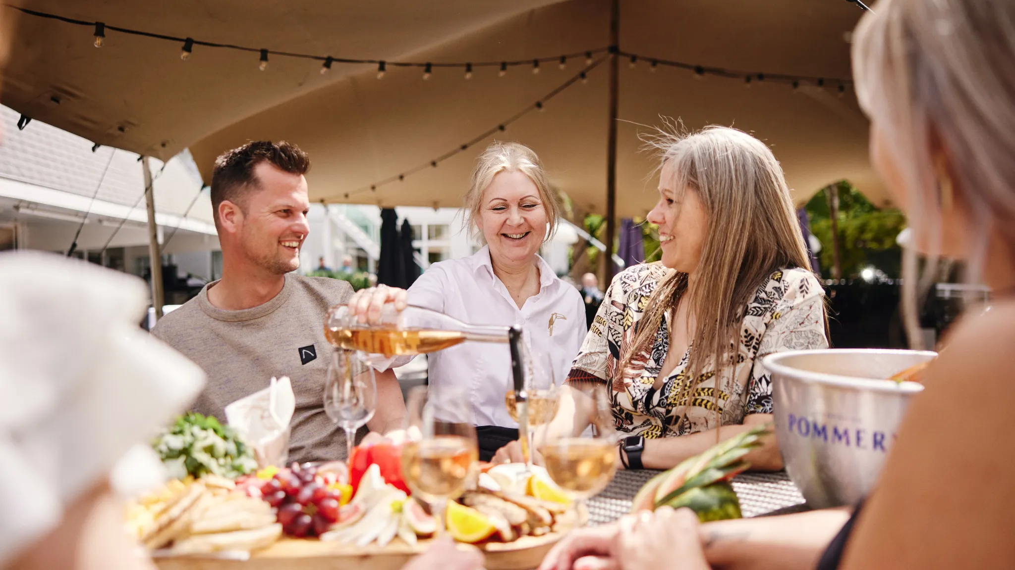 Eine Gruppe genießt gemeinsam eine Mahlzeit und die Gesellschaft der anderen unter einem großen Sonnenschirm auf der Terrasse.