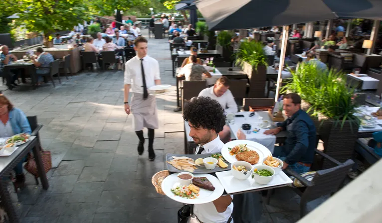 Auf der lebhaften und sonnigen Außenterrasse des Hotels serviert ein Kellner den Gästen Speisen.